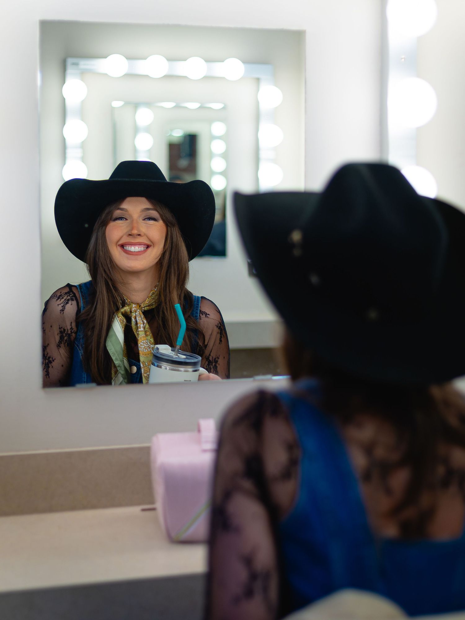 Woman in black cowboy hat smiling at dressing room mirror with Hollywood lights, holding a drink.