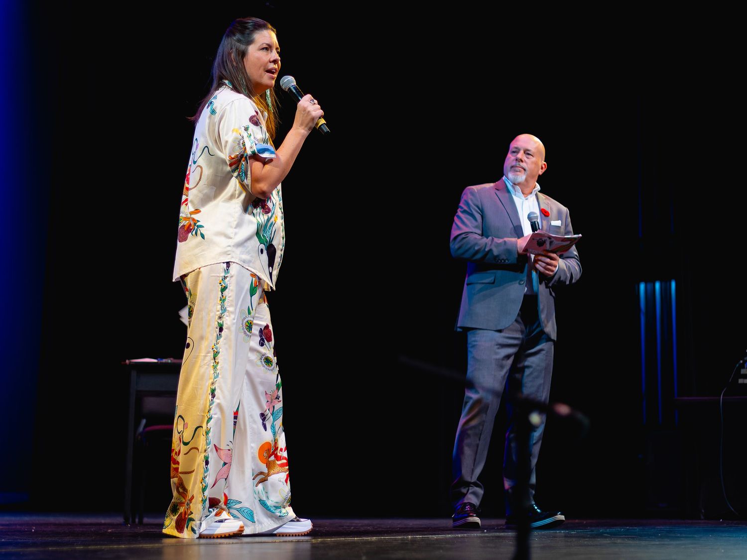 Two speakers on stage, woman in floral outfit holding microphone, man in gray suit at a formal event.