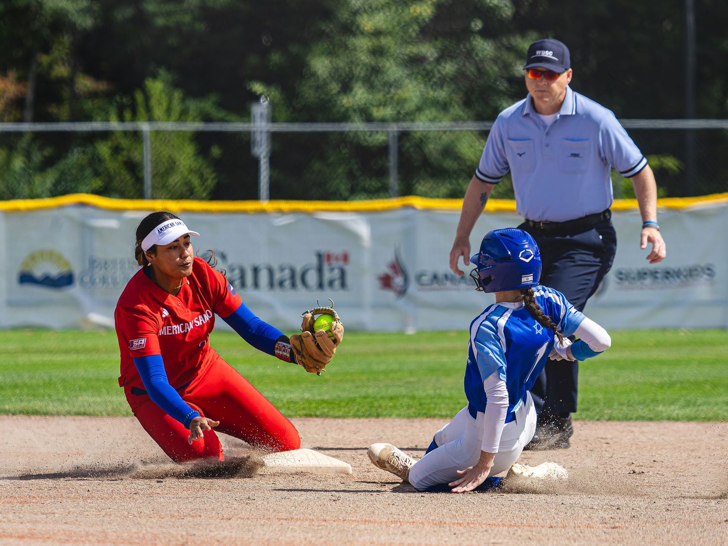 Softball player slides into second base as fielder catches ball, with umpire watching closely during a competitive game.