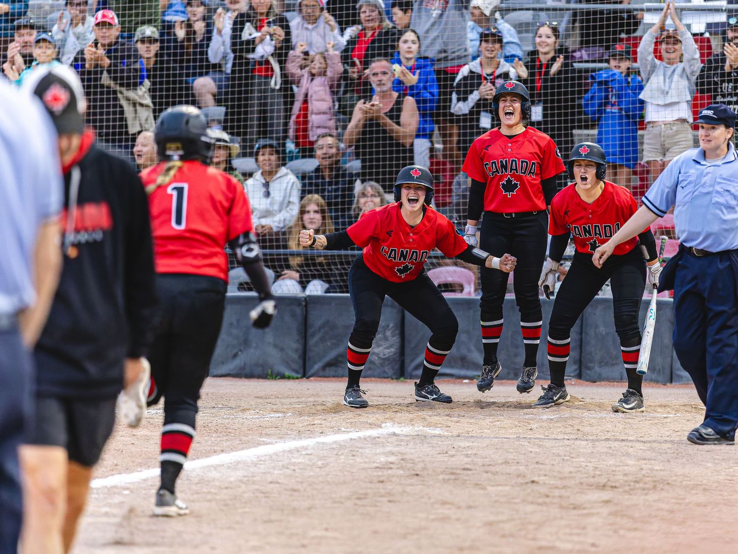 Softball players in red uniforms celebrate excitedly near home plate during a competitive game with fans cheering in background.