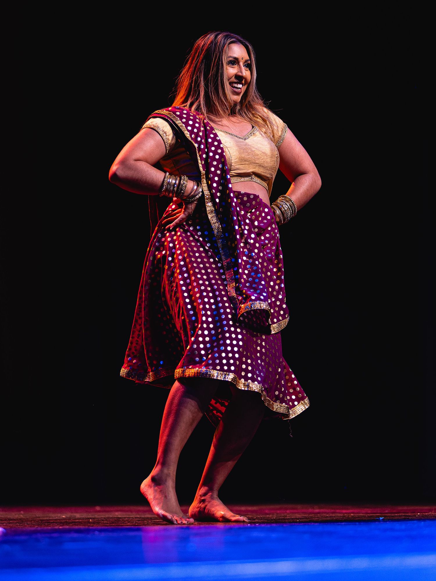 Woman in colorful Indian dance attire performing Bollywood dance on stage, smiling joyfully.