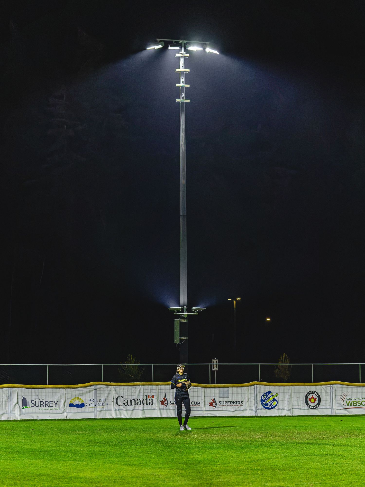 Athlete stands under bright stadium lights on green field at night with sponsor banners visible in background.