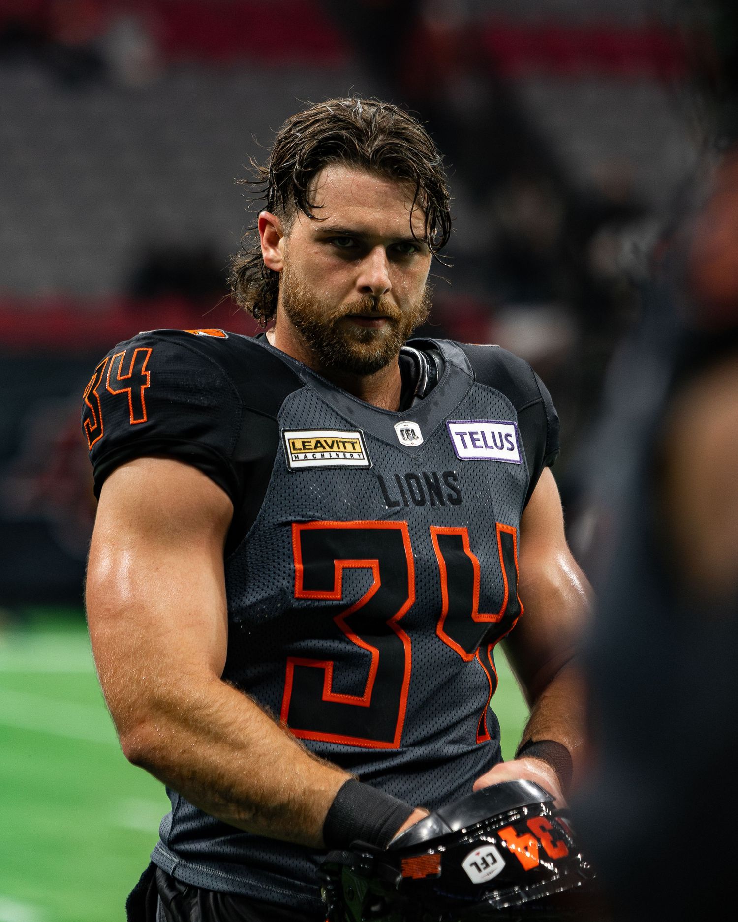 BC Lions player #34 in gray and orange uniform stands on field during CFL game at indoor stadium.