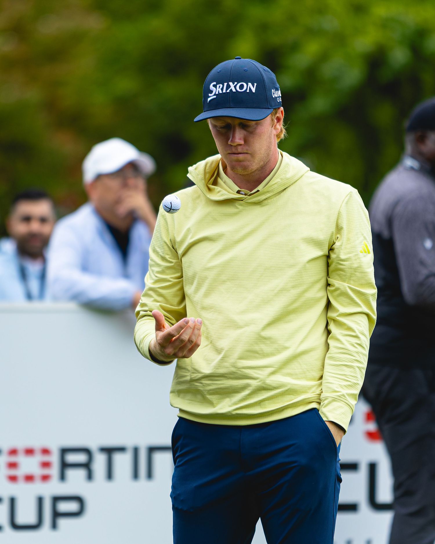 Golfer in yellow Srixon hoodie and navy pants stands on course, examining his hand near a tournament cup sign.