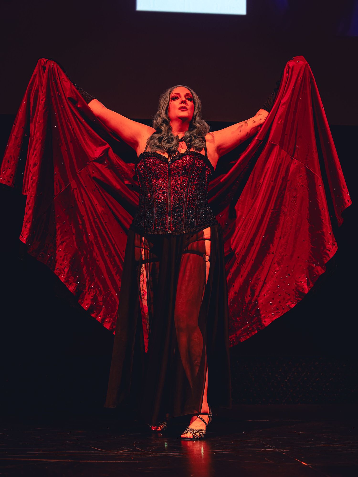 Performer in red sequined corset and dramatic crimson cape poses on stage under moody lighting.