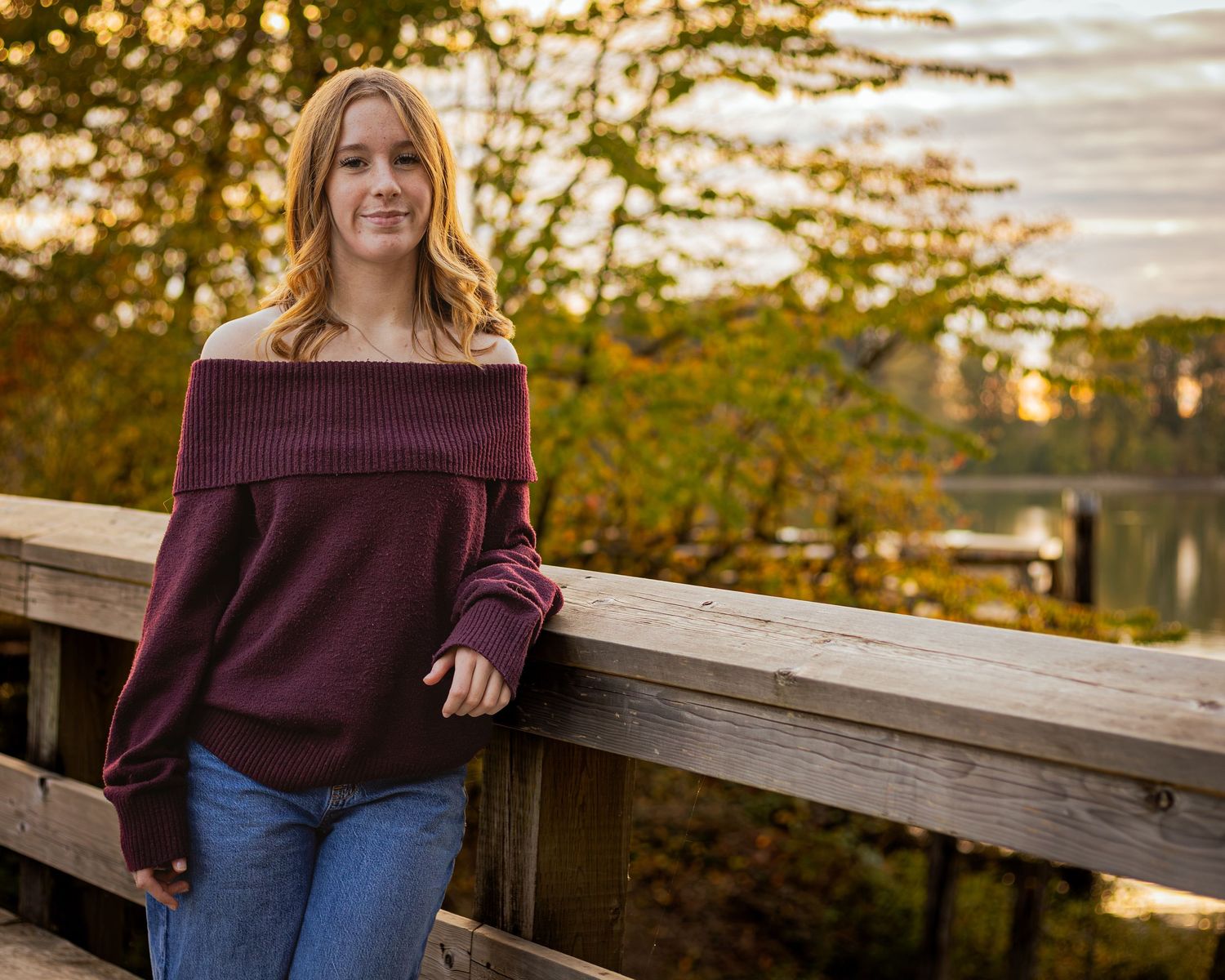 Young woman in burgundy off-shoulder sweater and jeans leaning on wooden bridge railing with autumn foliage backdrop.