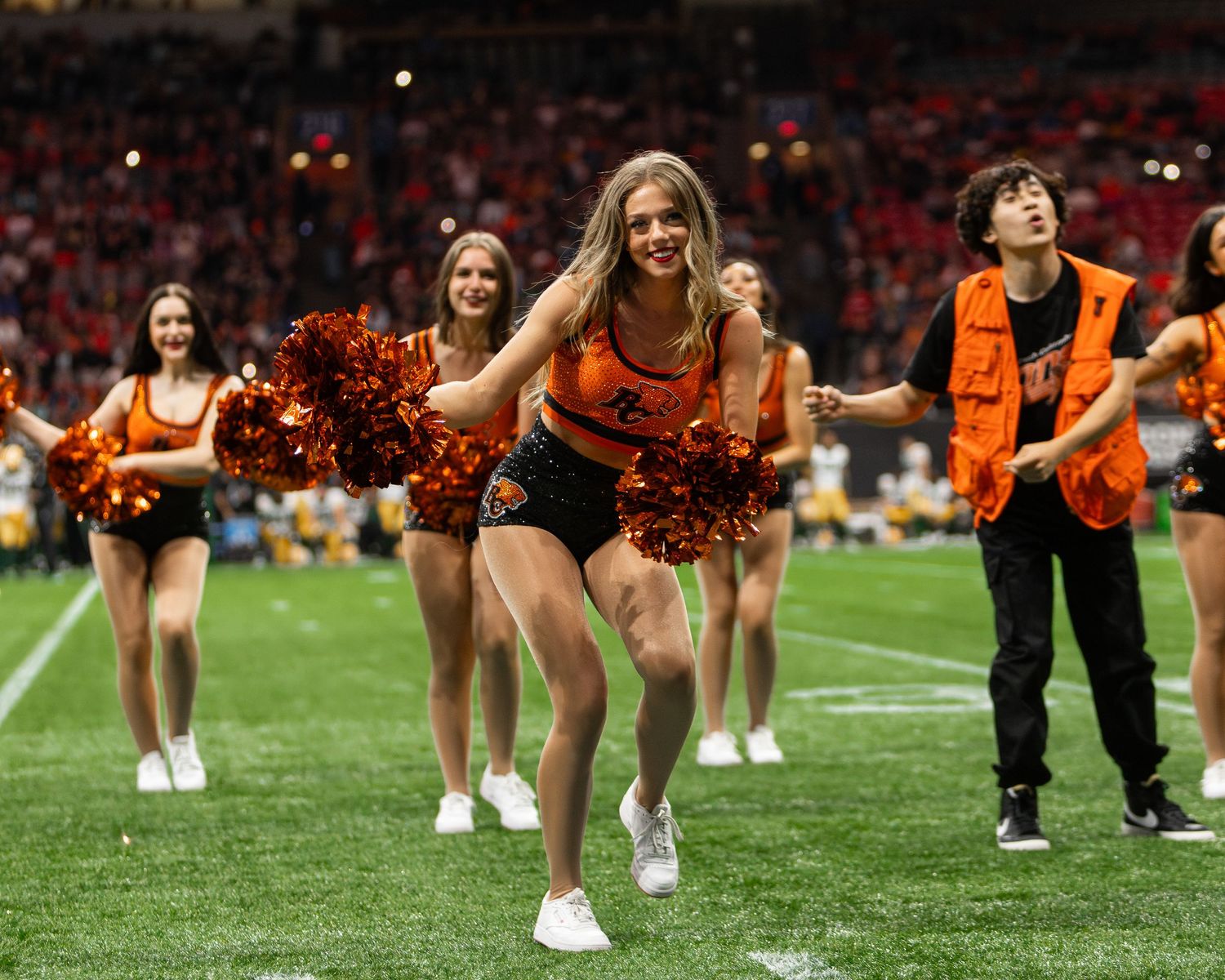 Cheerleaders in orange and black uniforms perform with pom-poms on a football field during a game.