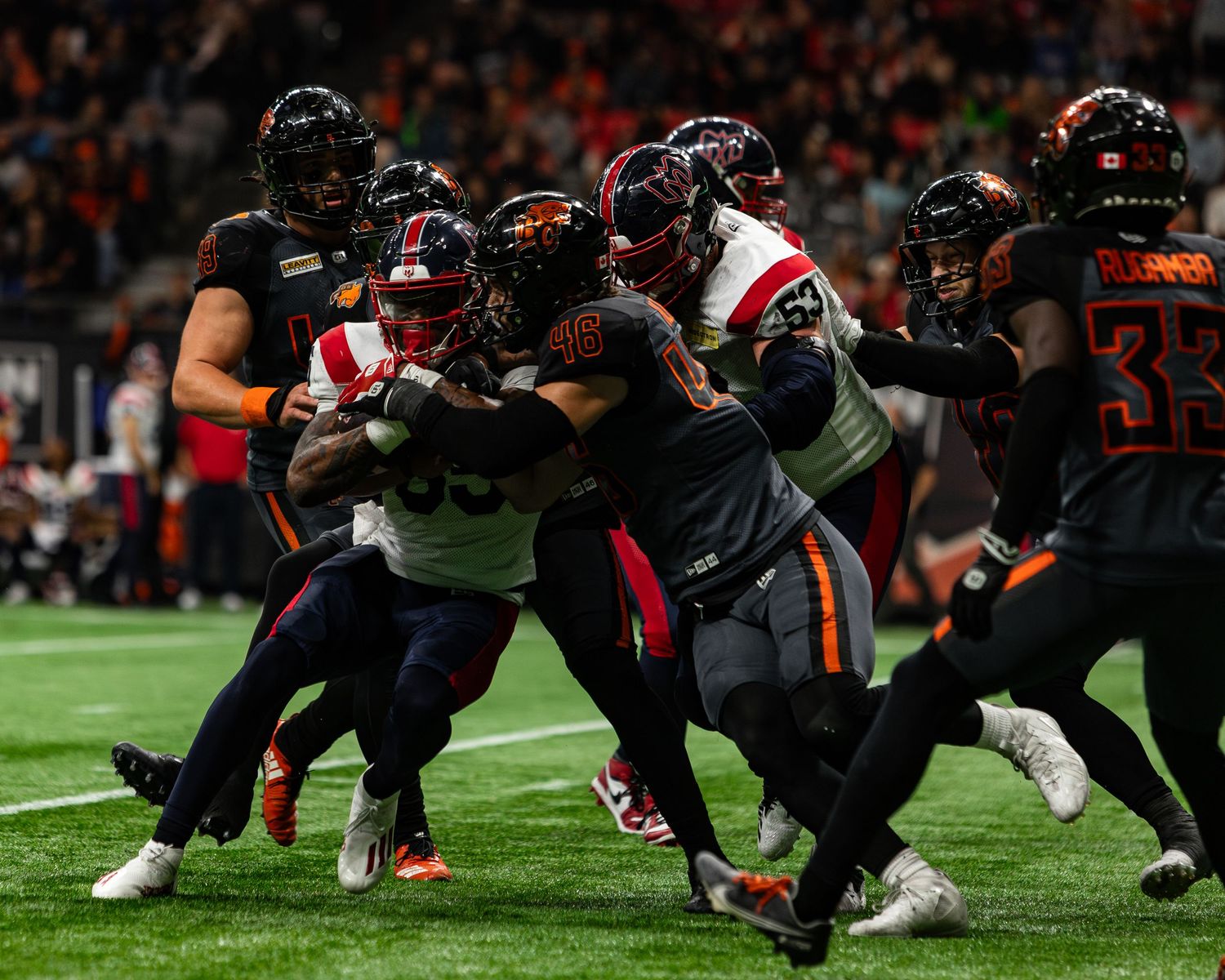 Football players clash in an intense CFL game tackle under stadium lights wearing red and black uniforms.