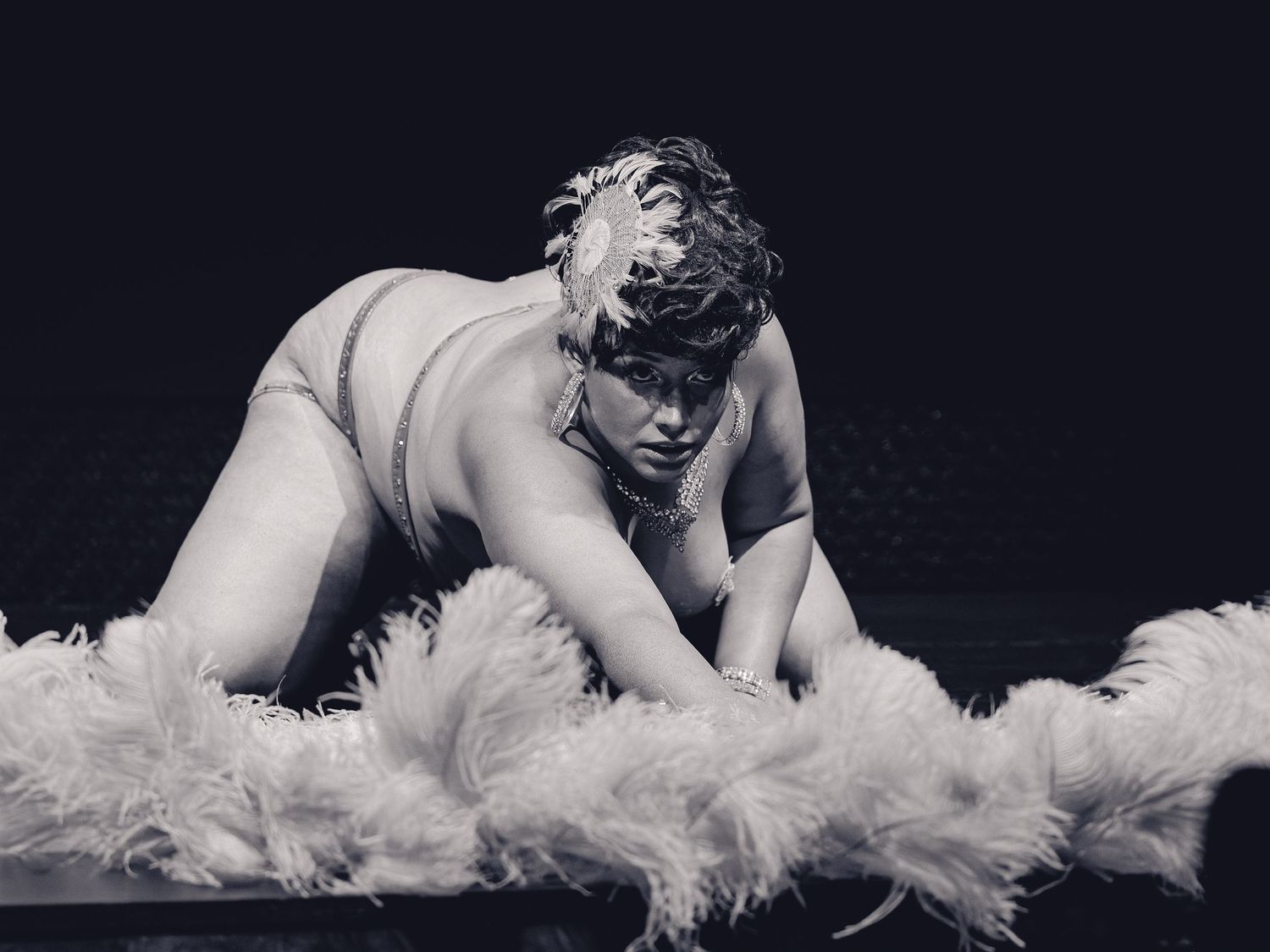 Black and white photo of a plus-size burlesque performer posing dramatically with white feather boa on stage.