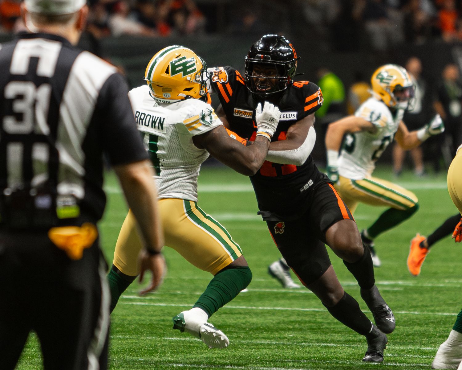 CFL players clash on field, BC Lions in orange vs Edmonton Elks in green uniforms during an intense indoor game.