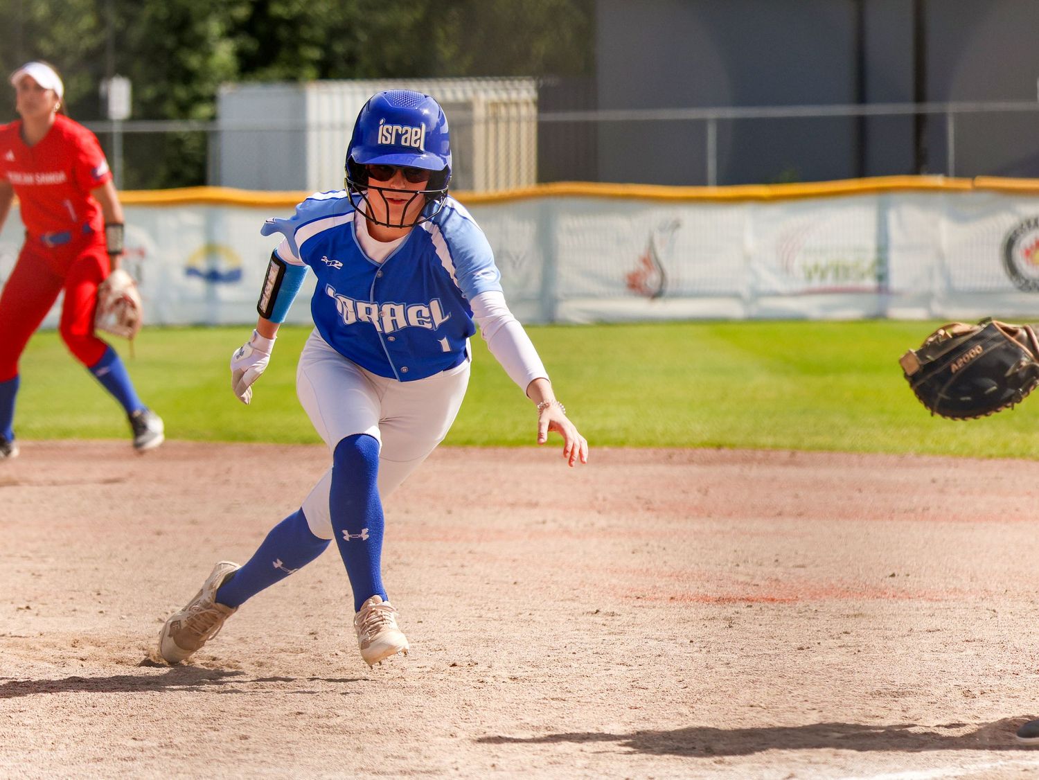 Female softball player in blue uniform rounding base during competitive game on sunny day.