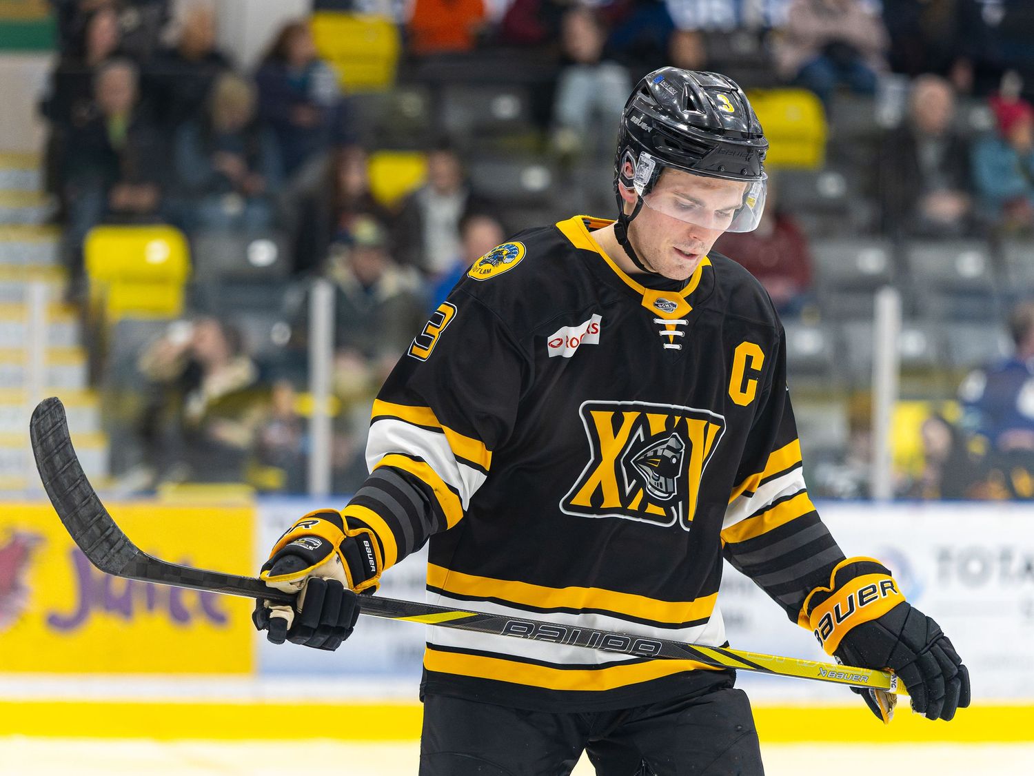 Hockey player in black and gold jersey with captain C patch skating on ice rink during a game.