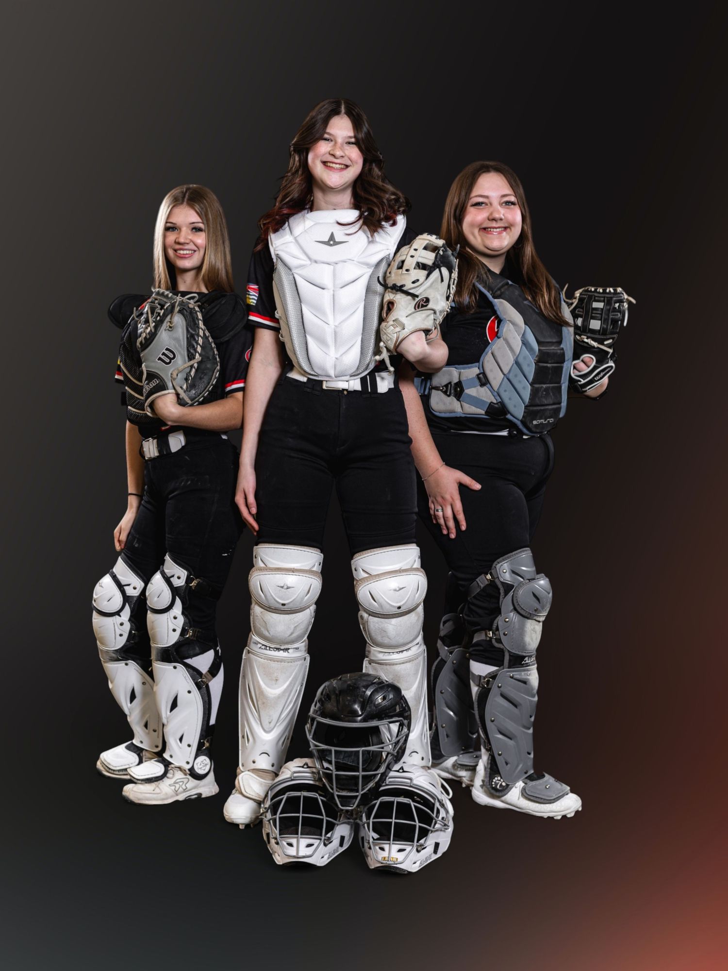 Three girls in softball catcher gear posing together, wearing chest protectors, shin guards, and holding mitts.