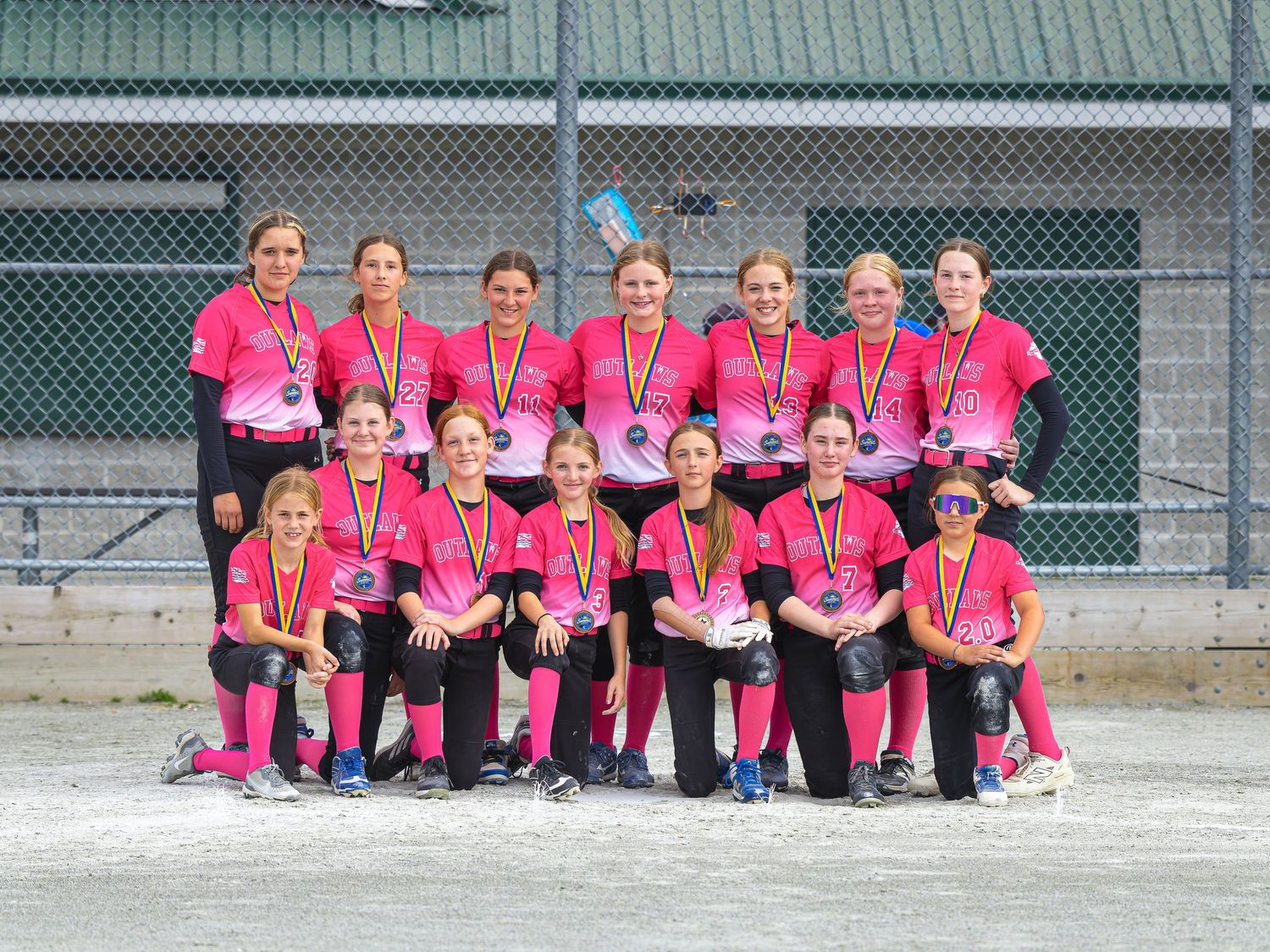 Youth girls softball team in pink uniforms posing for group photo on dirt field, wearing medals after tournament win.