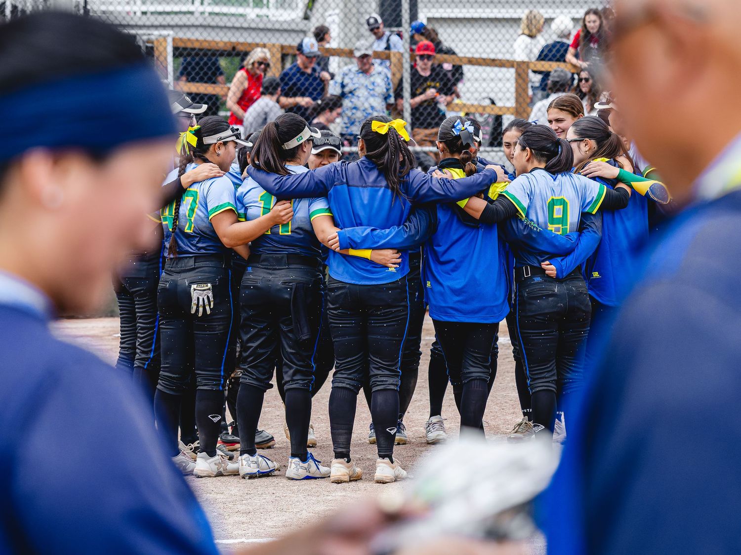 Softball team huddles together in blue and black uniforms during a game, arms around each other on the field.