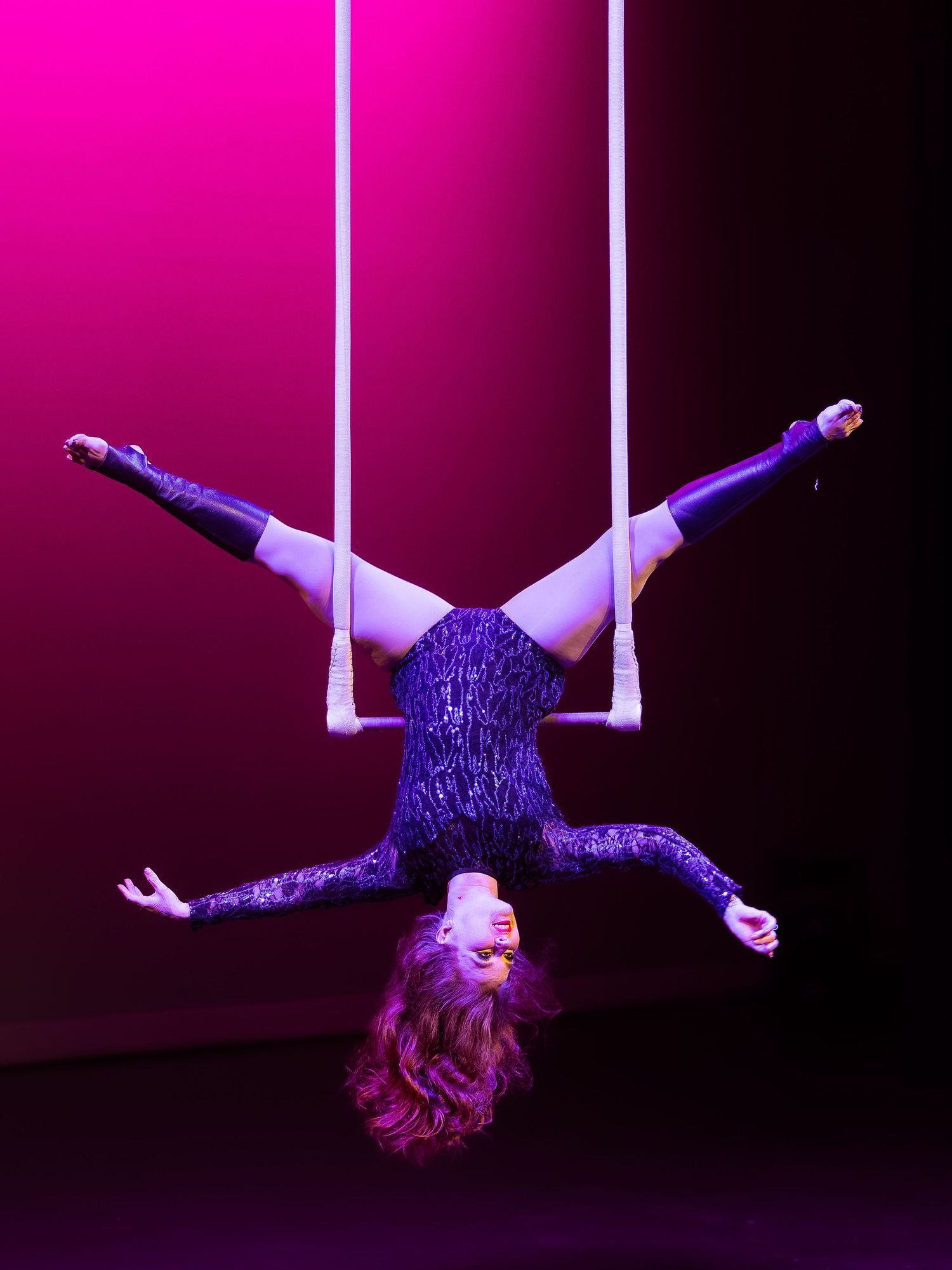 Aerial acrobat in purple sequin outfit performs inverted split pose on trapeze against pink-lit backdrop.