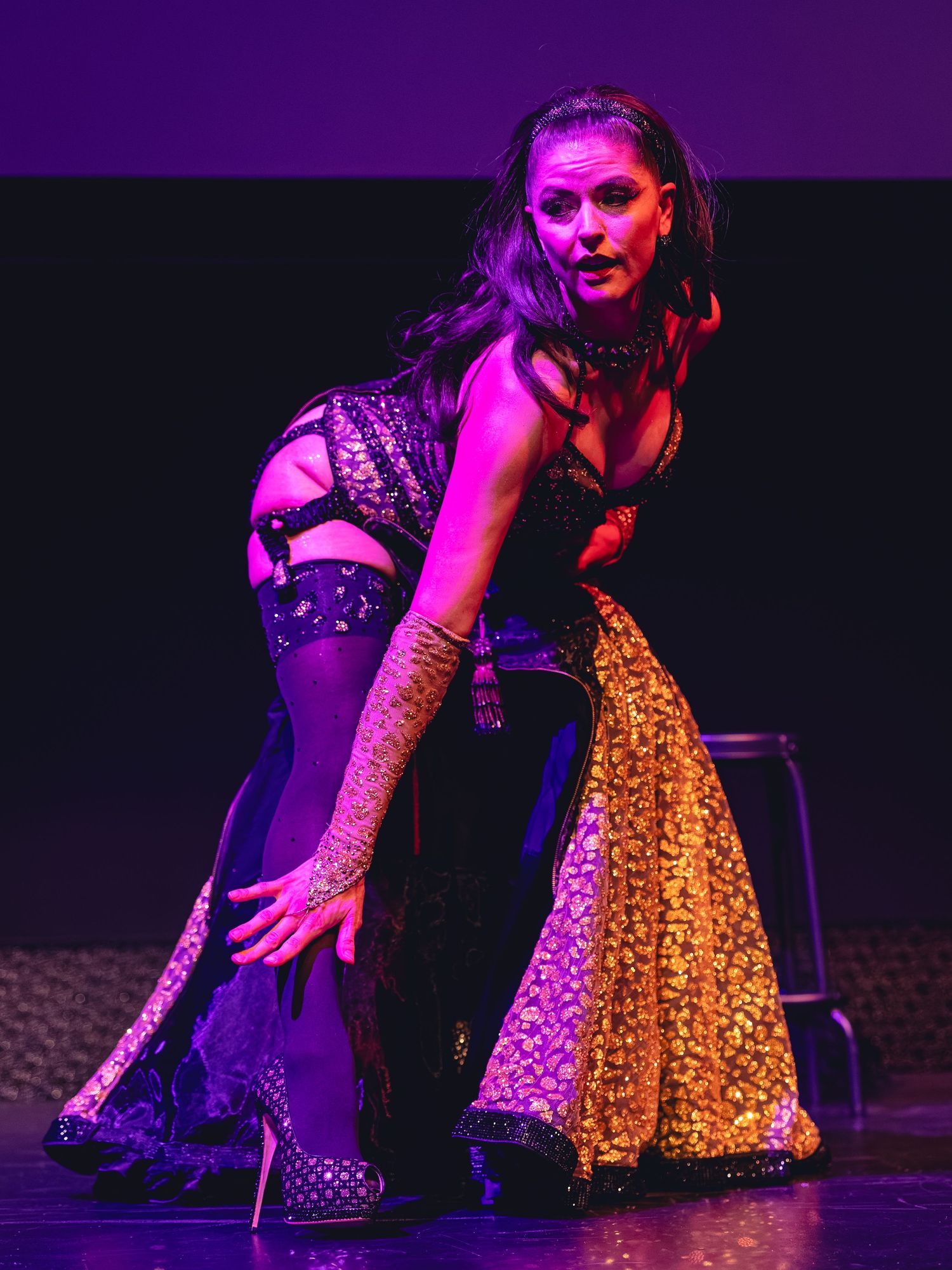Performer in elaborate costume with gold sequined skirt and black lingerie poses dramatically on stage under purple lighting.