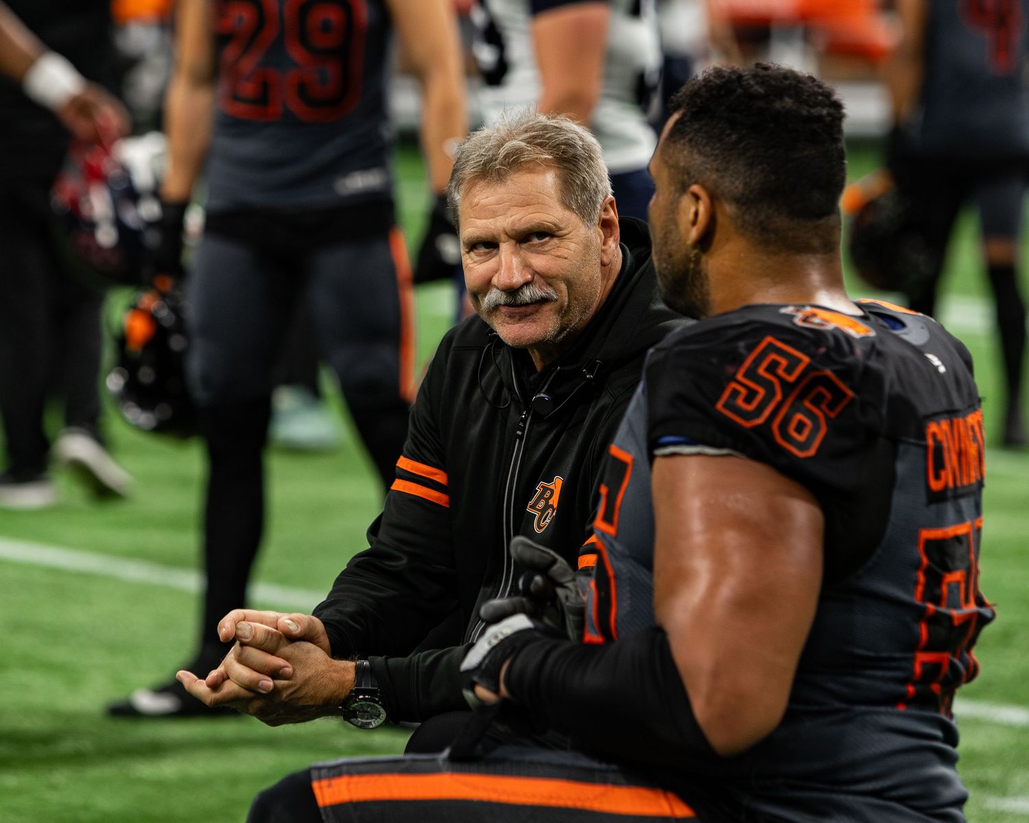 Coach and player #56 share a conversation on the sideline during a football game in orange and black uniforms.