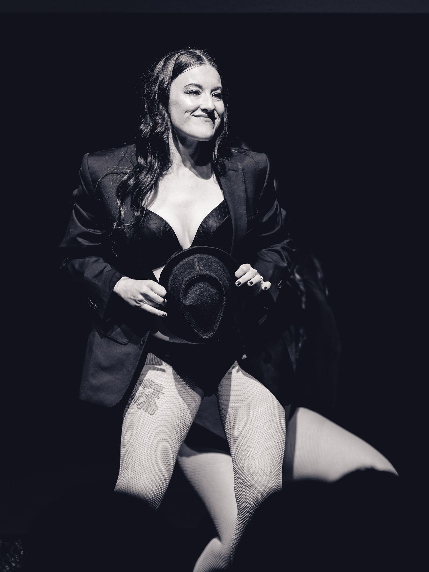 Black and white photo of a woman performing on stage in a bodysuit and blazer, holding a hat.