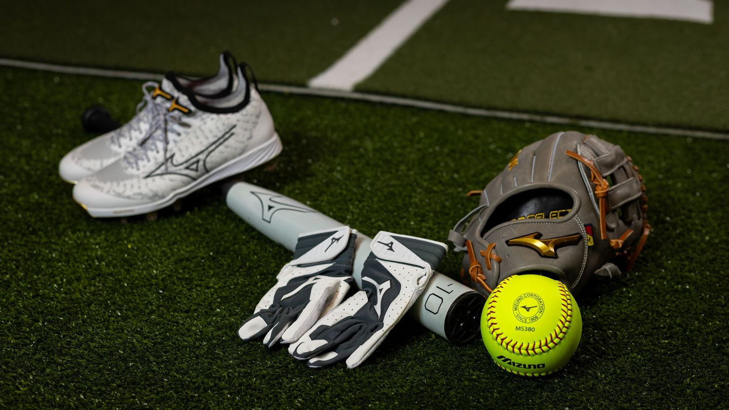 Softball equipment on green turf including cleats, batting gloves, a fielding glove, and a yellow softball.
