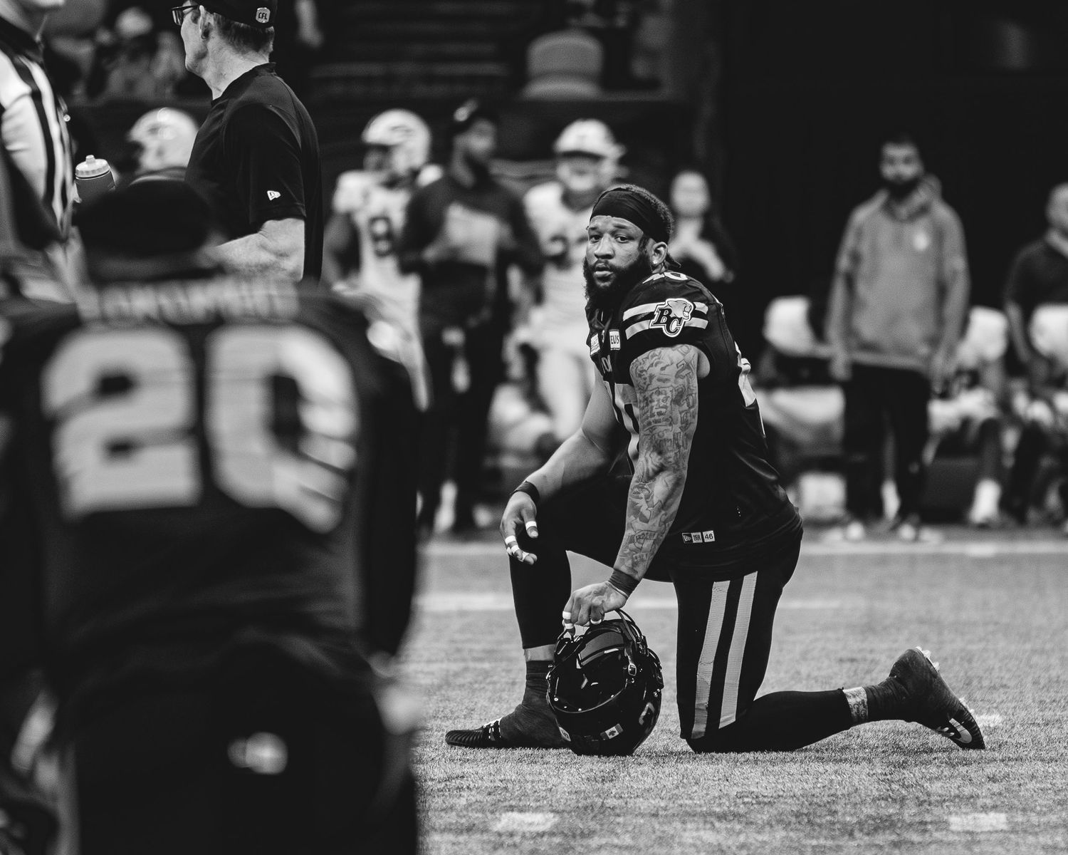 Black and white photo of an NFL player kneeling on the field holding his helmet during a football game.