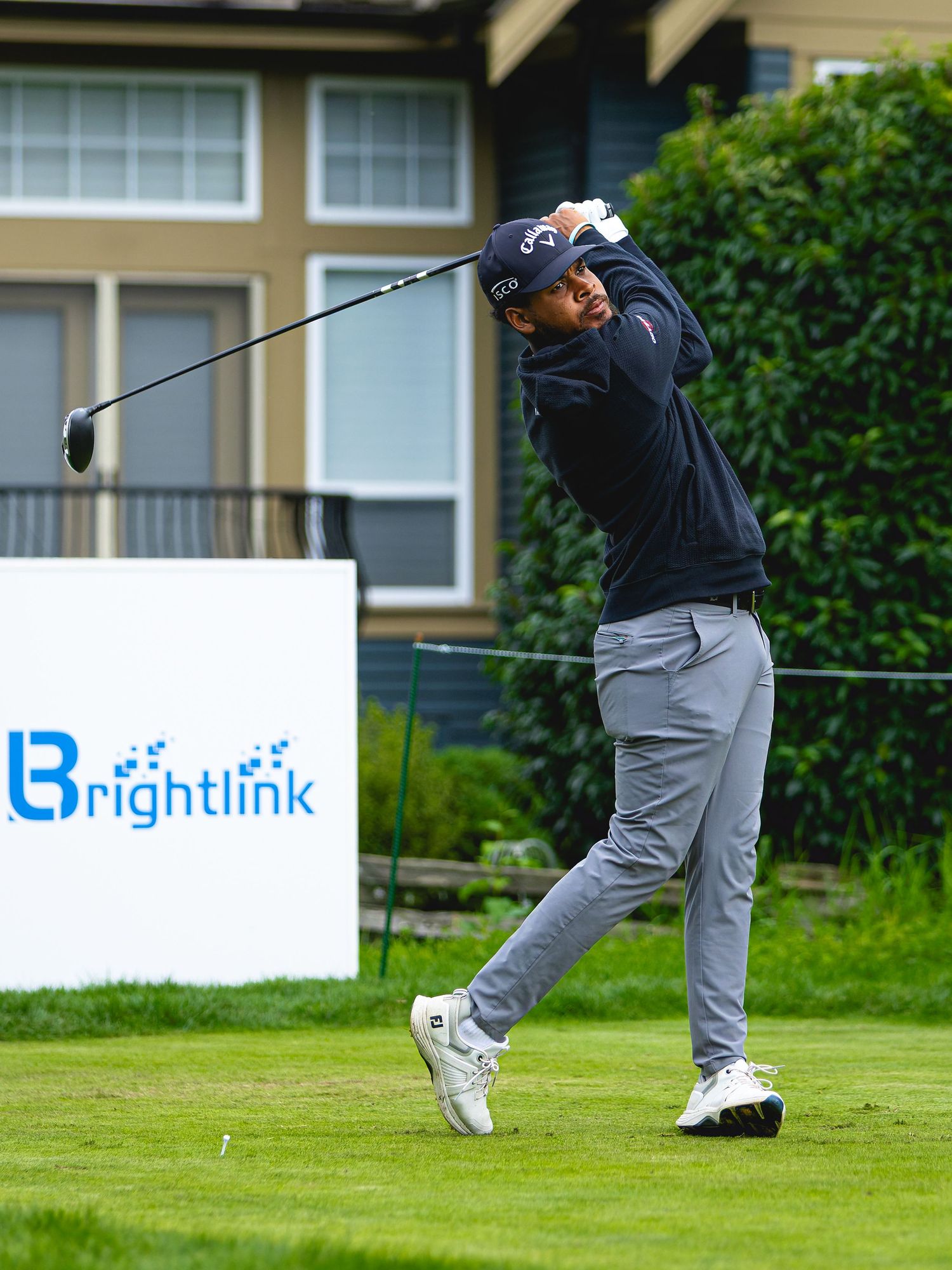 Golfer in navy Callaway attire swings a driver at a Brightlink-sponsored golf tournament on a lush green course.