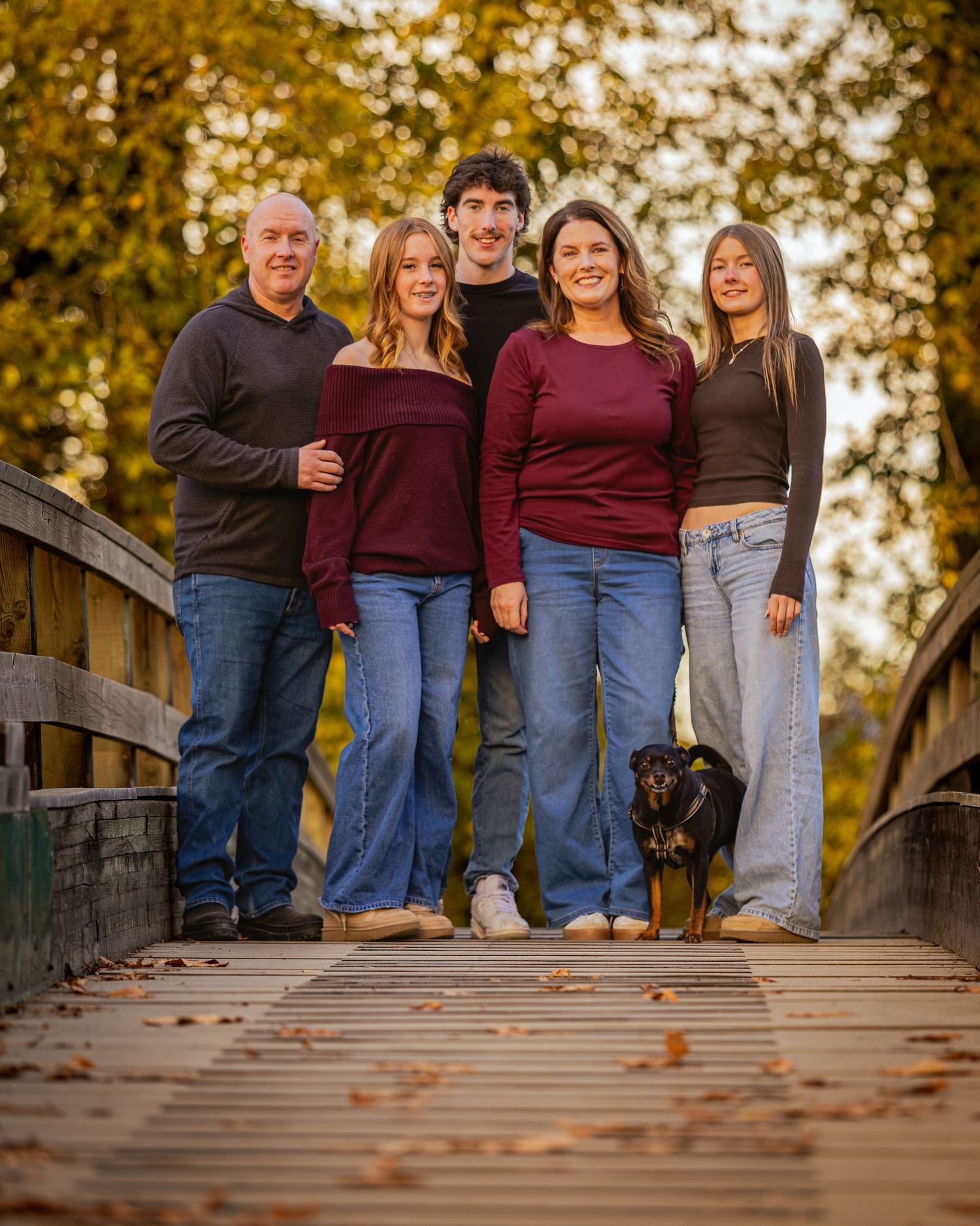 Family of five posing on a wooden bridge surrounded by autumn foliage with a small black dog.