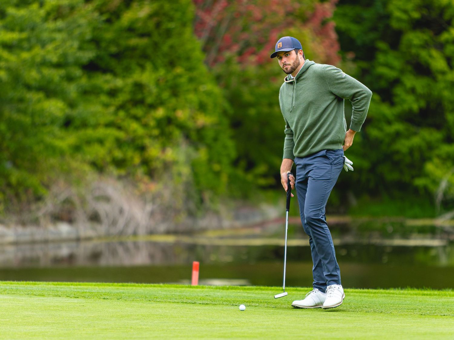 Golfer in green pullover and navy cap reads a putt on a lush green course with trees and water behind.