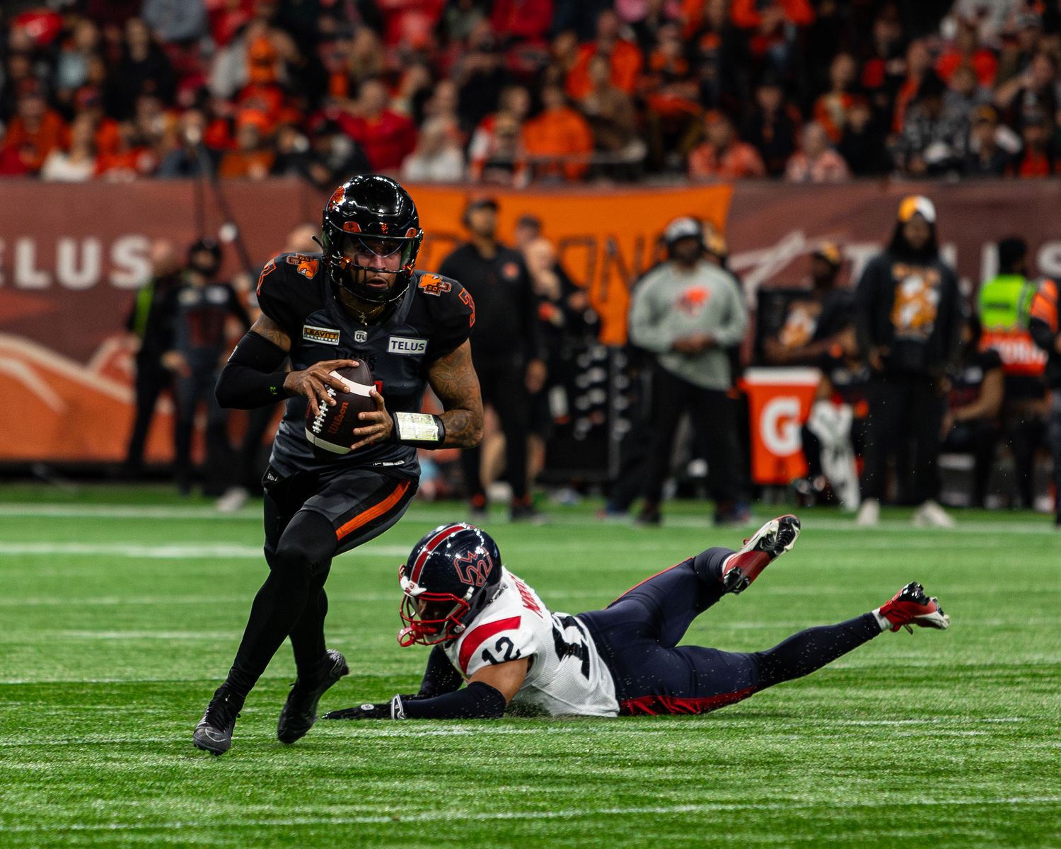 CFL quarterback in black uniform evades diving defender in navy jersey during indoor football game.