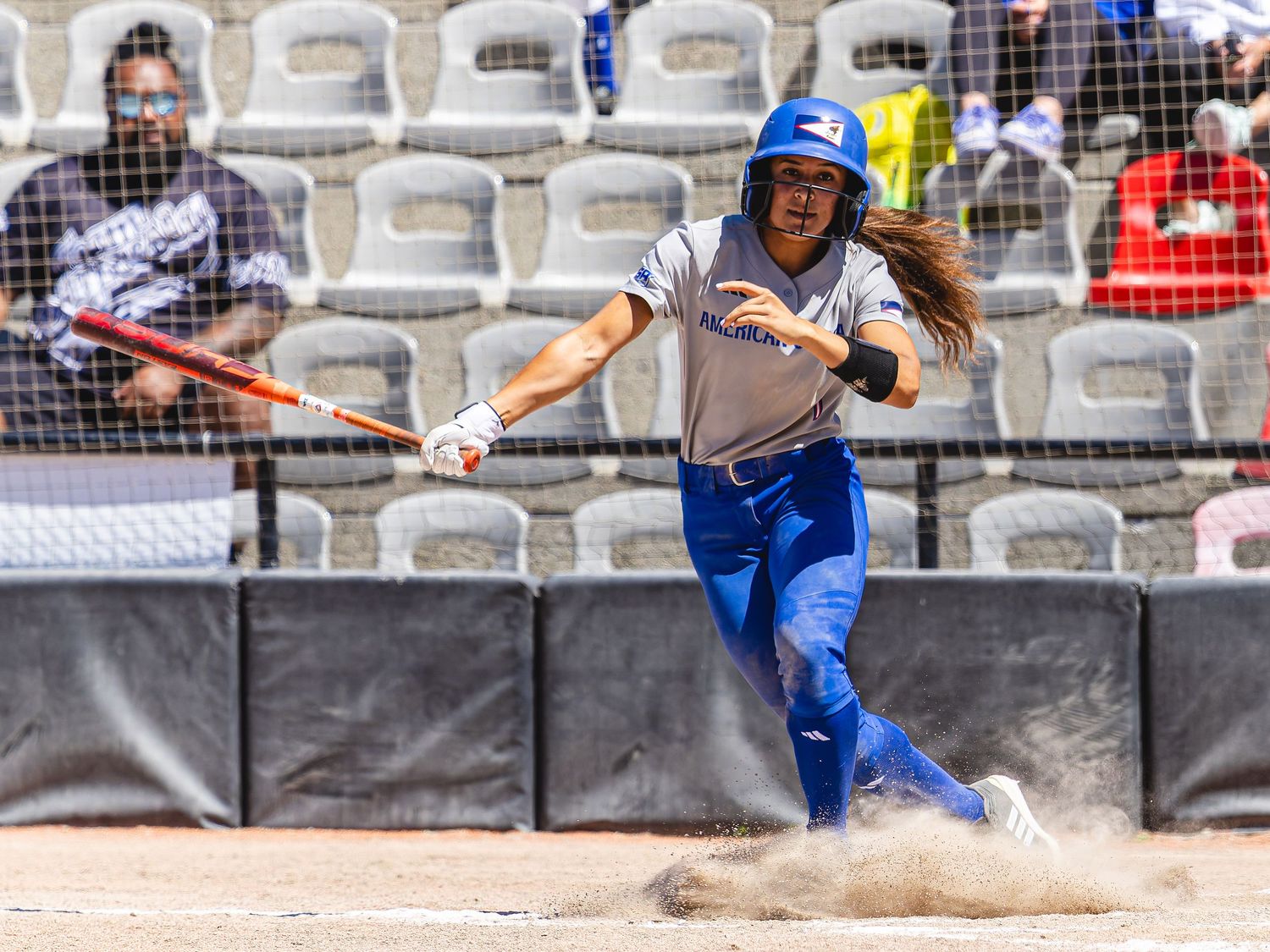 Female softball player in blue uniform slides into base during a sunny game, bat in hand, dirt flying.