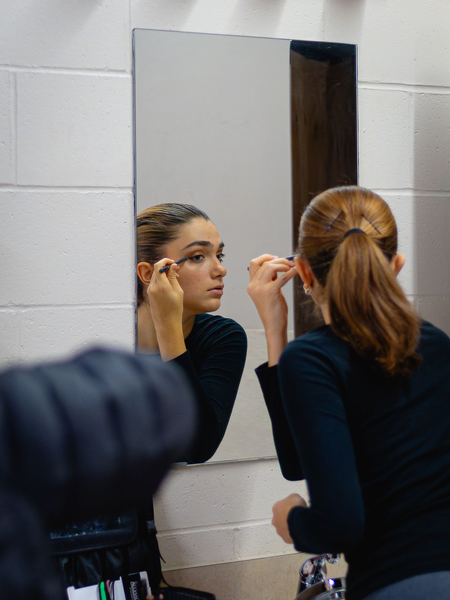 A young woman applies mascara while looking in a mirror in a dressing room before a performance.