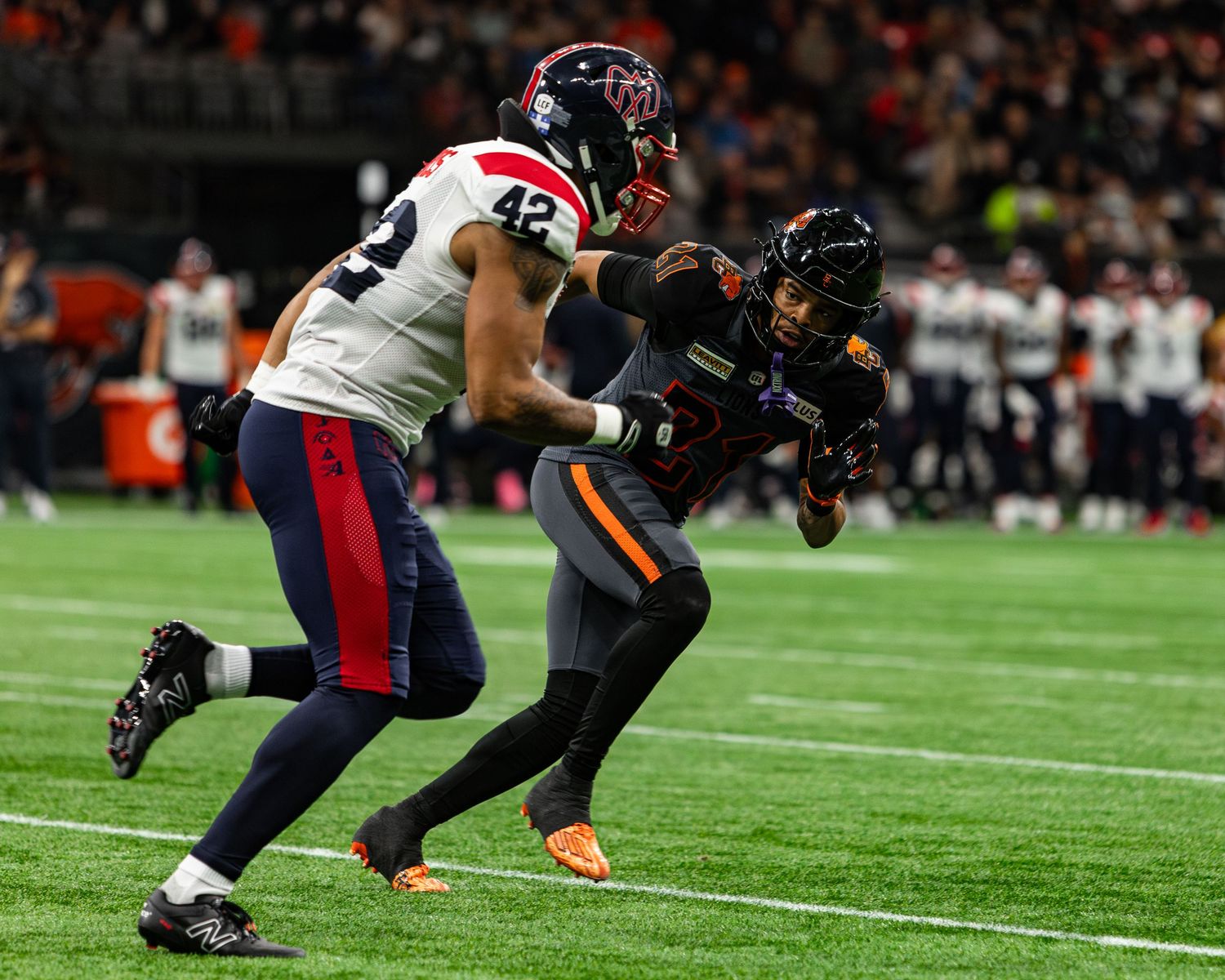 Football player in orange uniform evades tackle from defender wearing number 42 white jersey during indoor game.