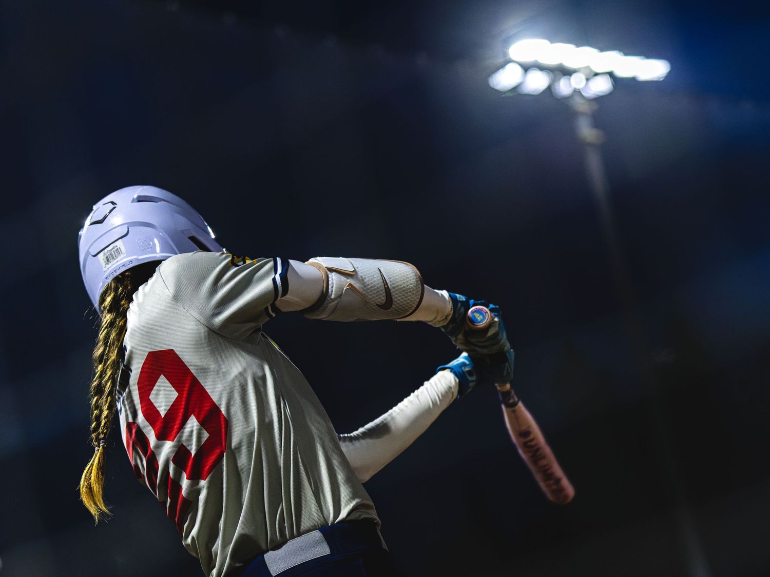 softball batter with braids and white helmet swings under bright stadium lights at night game.