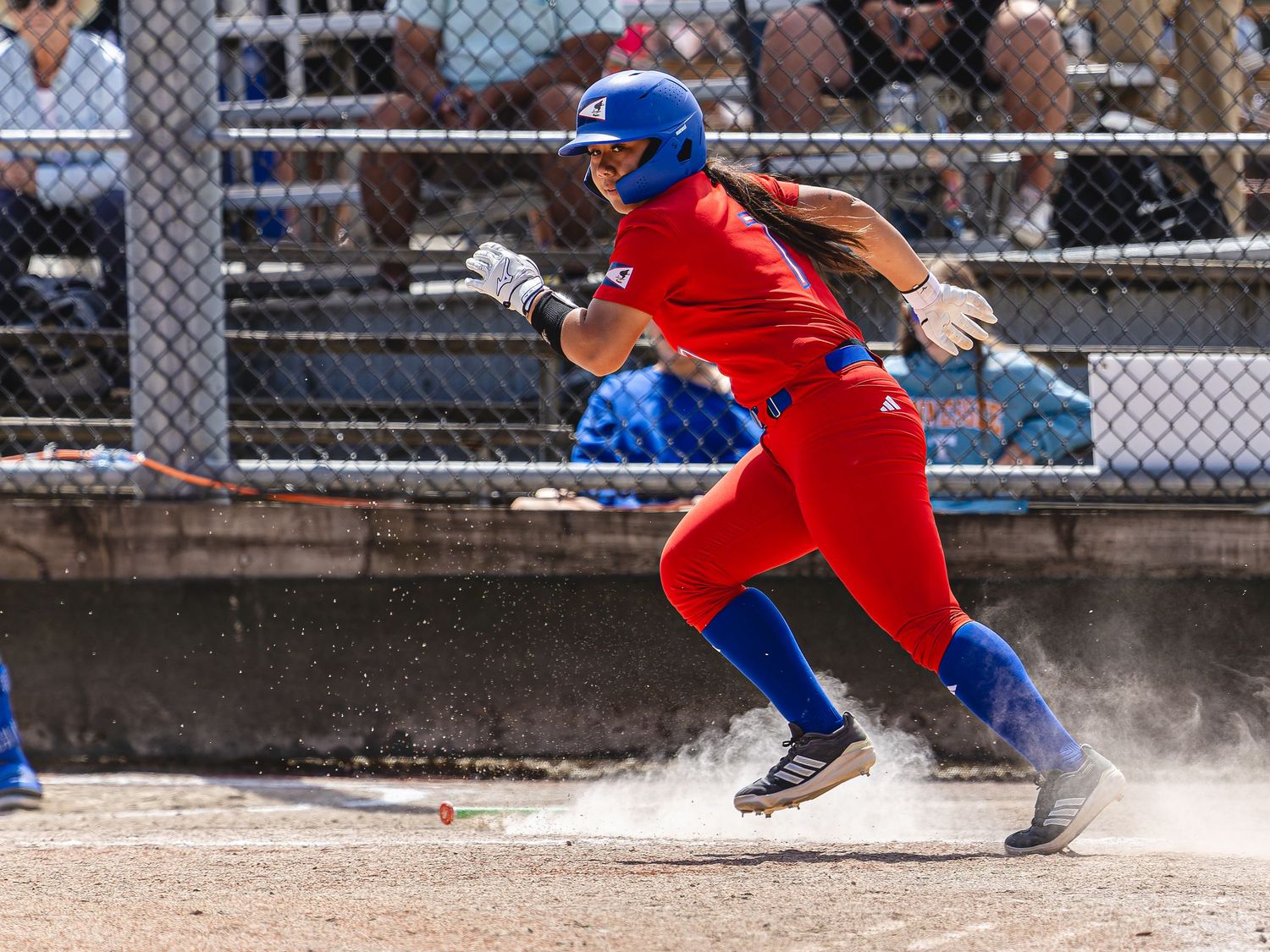 Female softball player in red and blue uniform sprinting toward base in a dusty outdoor game.