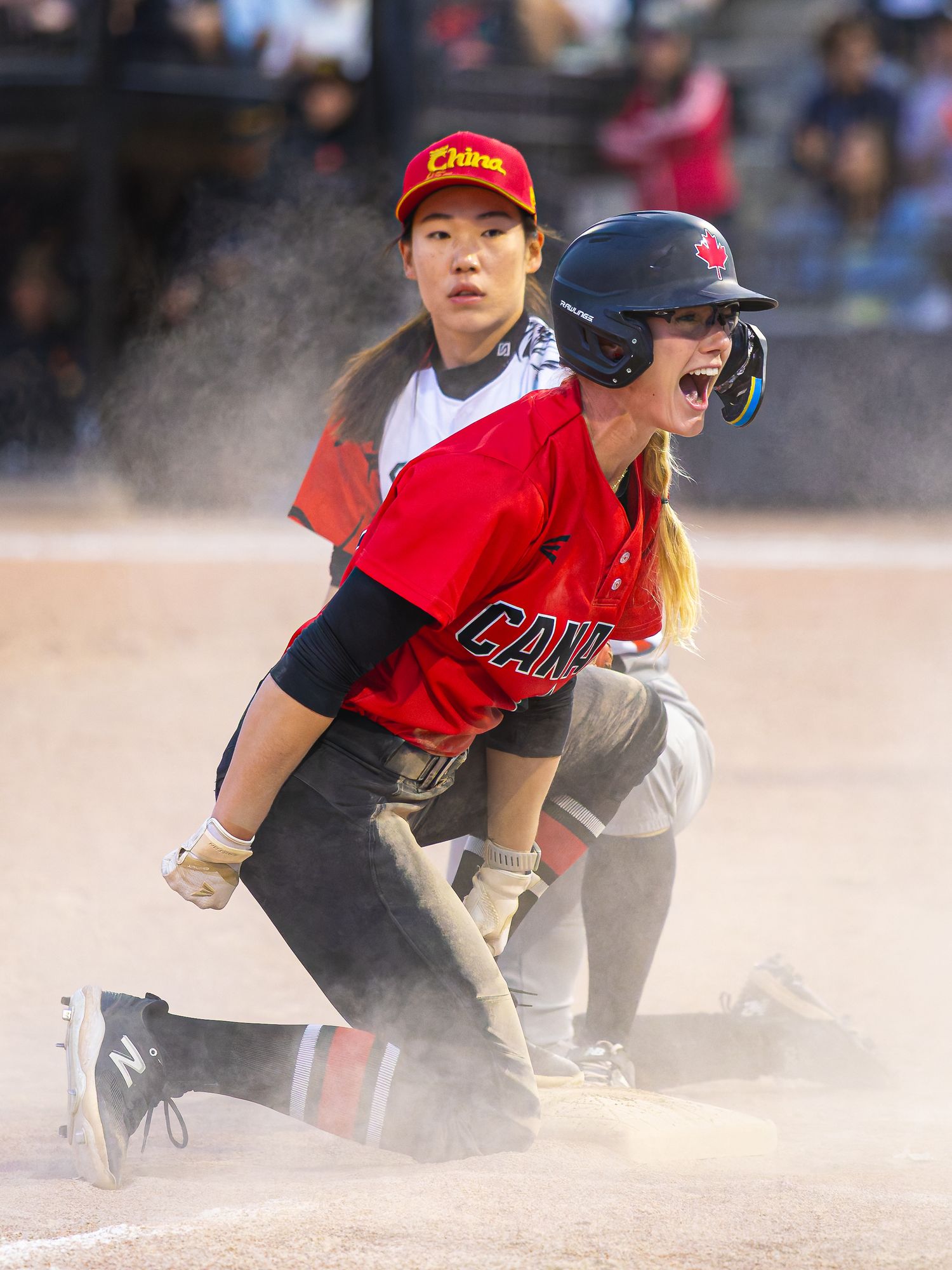 Canada softball player slides into base in cloud of dust as China fielder looks on during international game.