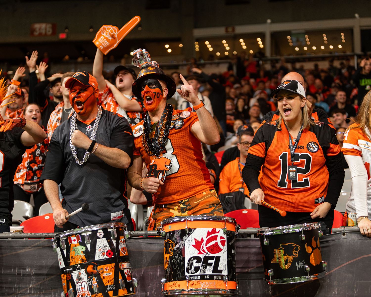 Excited CFL fans in orange and black face paint cheer loudly at a BC Lions football game.