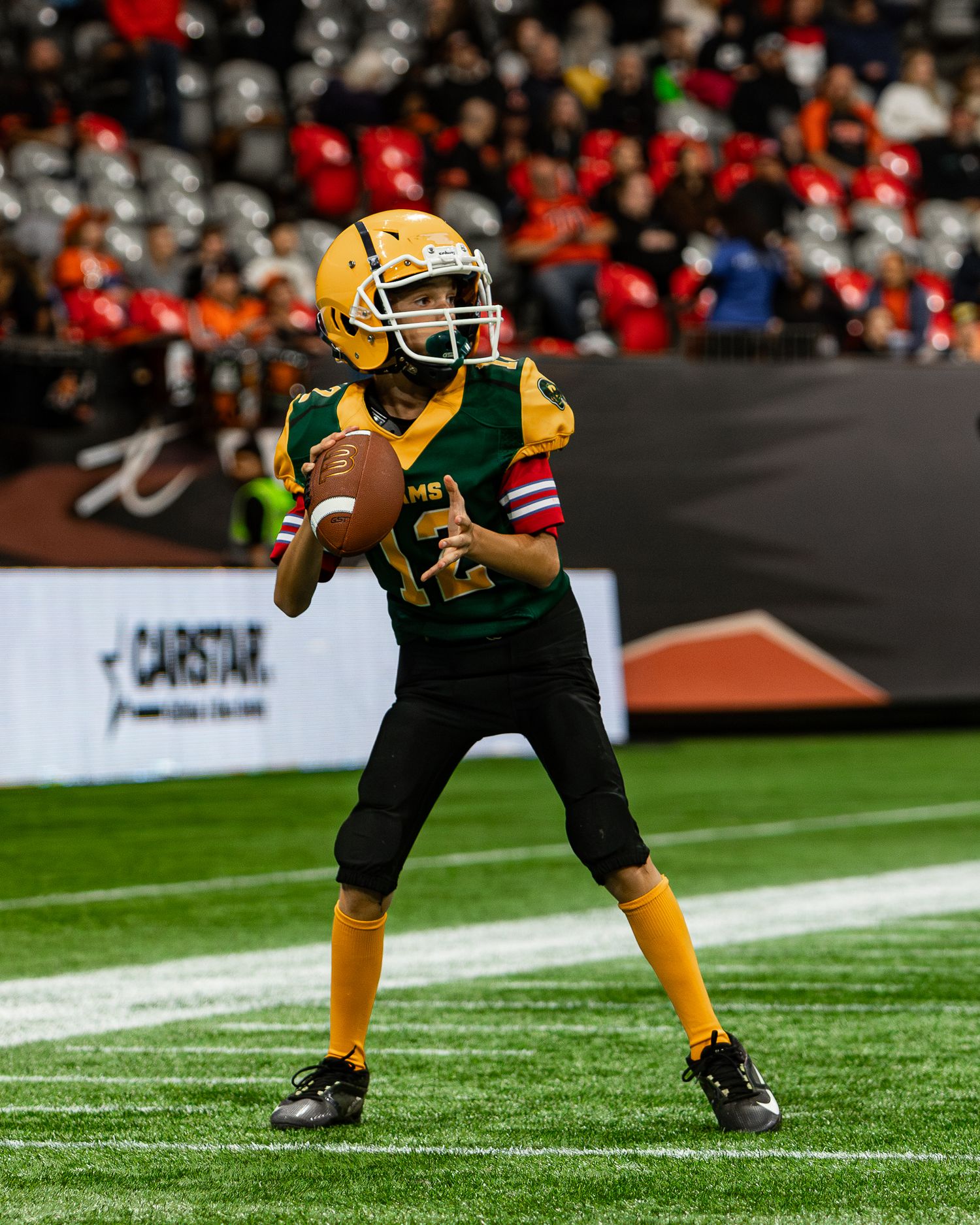 Young football player in green and gold uniform prepares to throw a pass during an indoor youth football game.