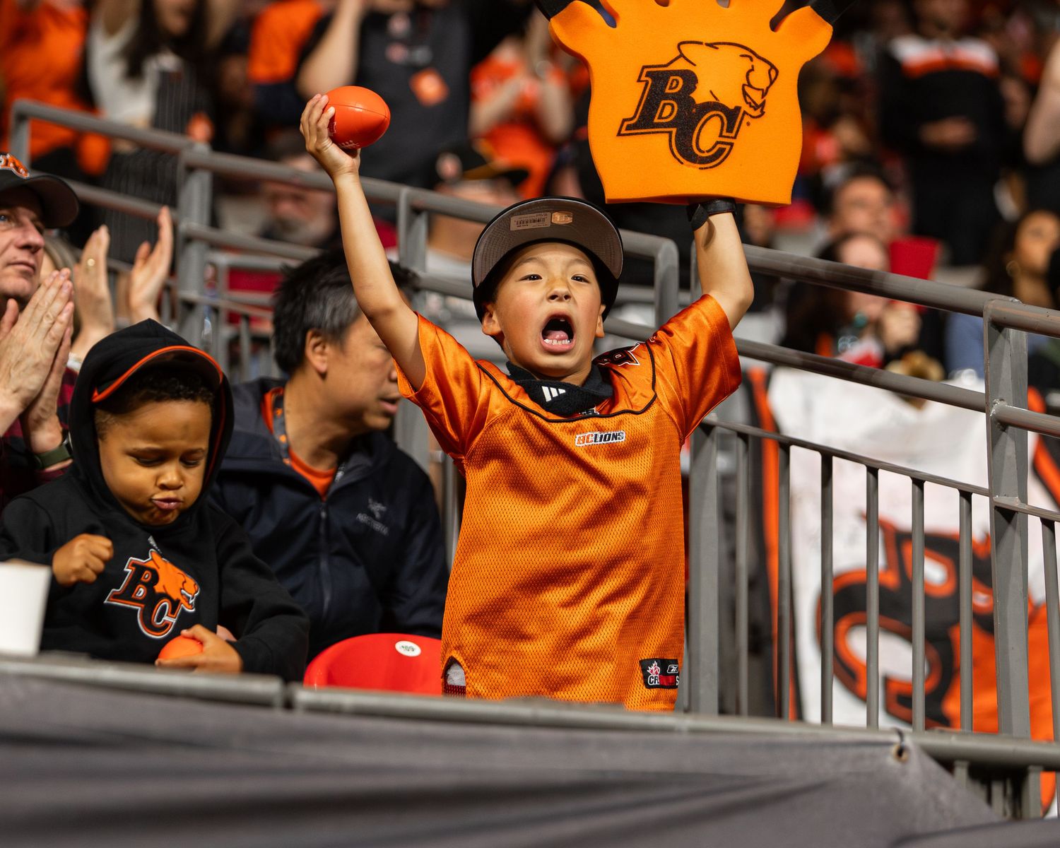 Excited young BC Lions fan cheering in stands, wearing orange jersey, holding team sign at CFL game.