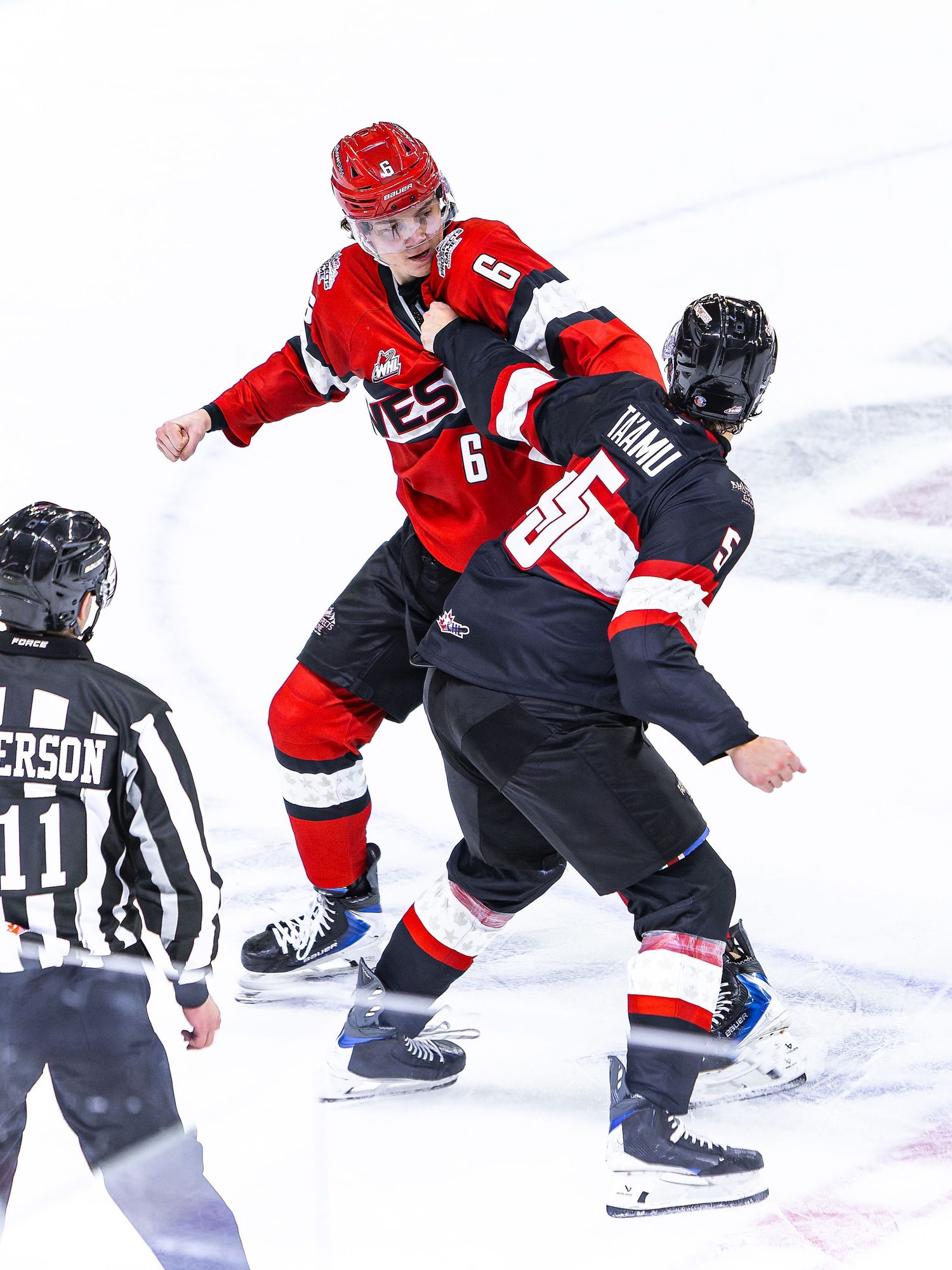 Two ice hockey players in red and black uniforms engage in a fight on the ice while a referee watches nearby.