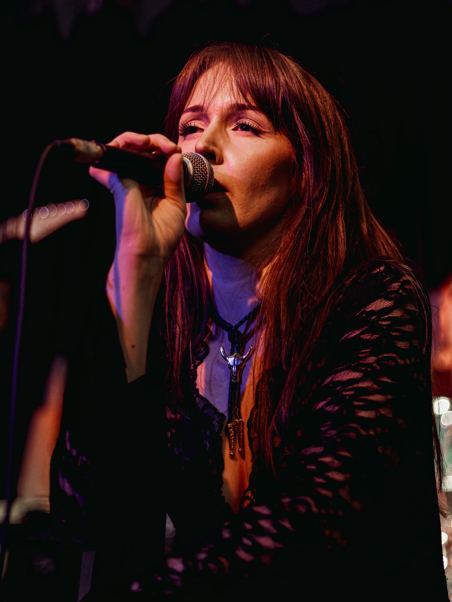 Female singer with long brown hair performing live on stage, holding microphone in black lace top under purple stage lighting.
