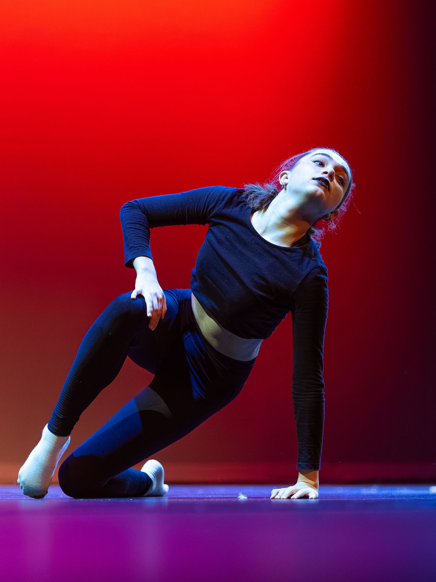 A female dancer in black activewear performs an expressive floor routine on stage with dramatic red lighting.