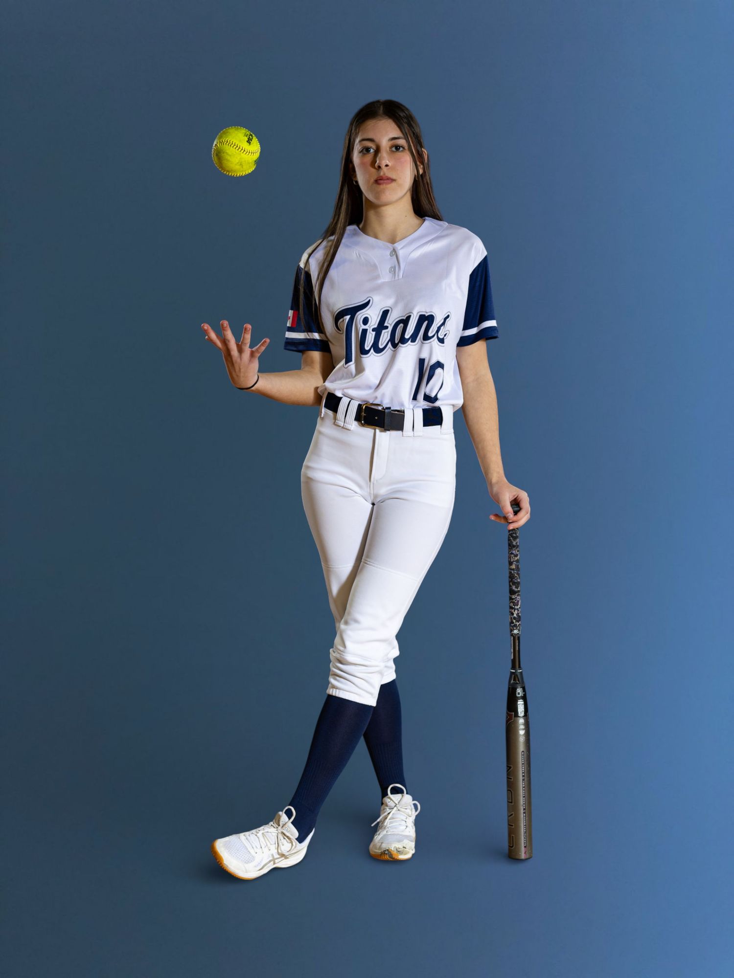 Female softball player in Titans uniform tossing a yellow ball, holding a bat, wearing white pants and navy socks.