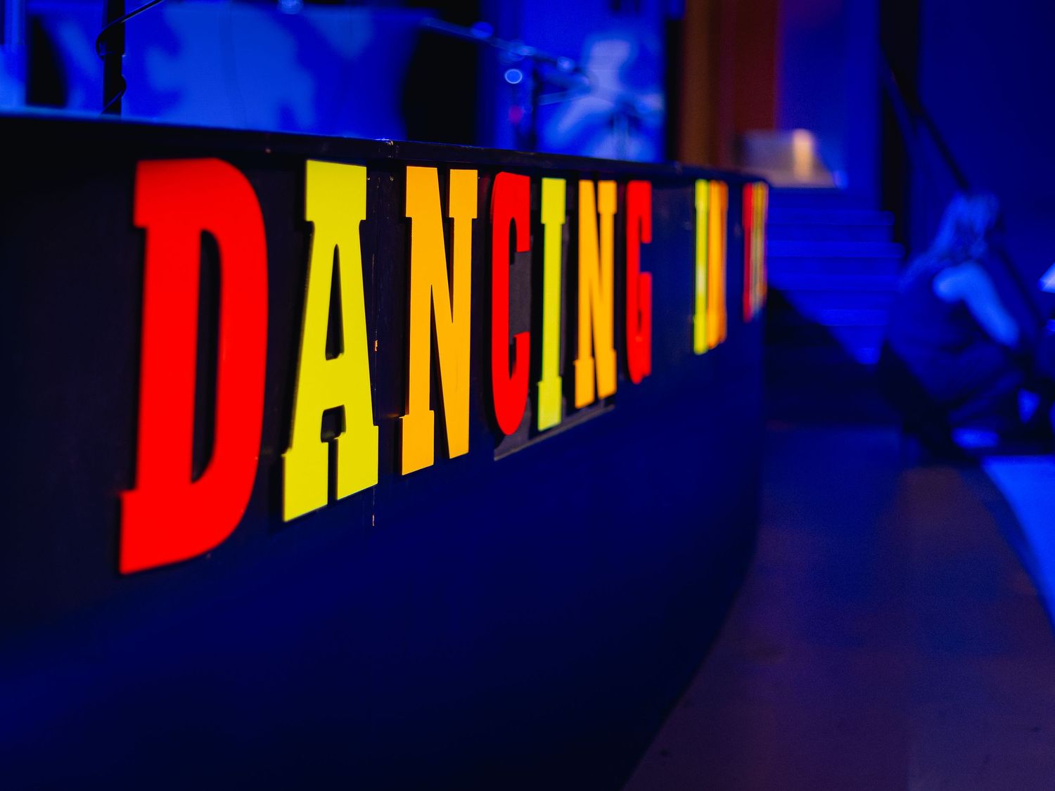 Illuminated DANCING sign with bold red and yellow letters glowing against a dark blue nightclub background.