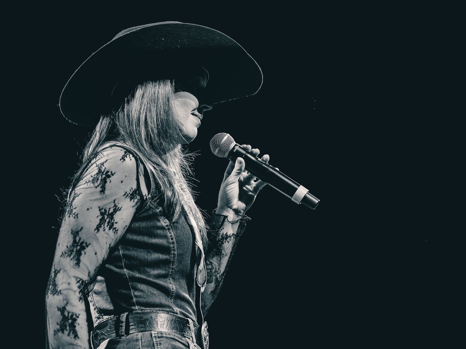 Black and white photo of a female country singer performing on stage wearing a wide-brim hat and holding a microphone.