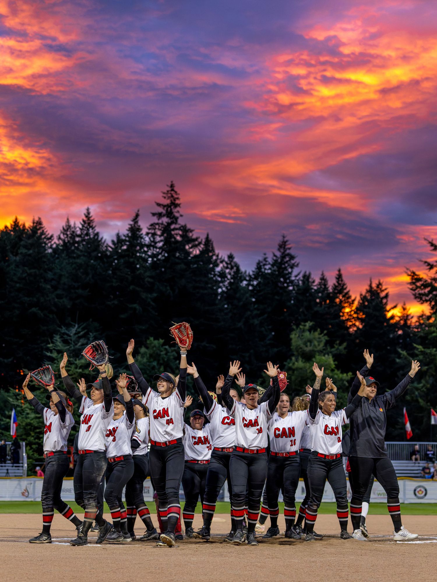 Canada softball team celebrates victory at sunset with vibrant orange and purple sky backdrop on the field.