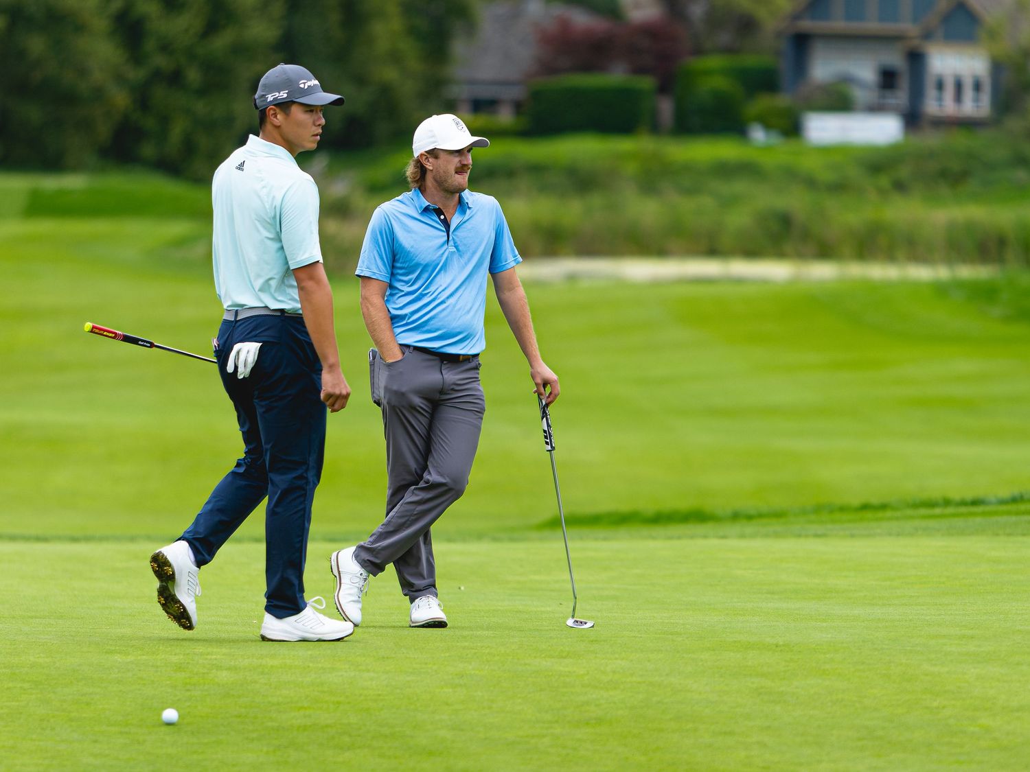 Two golfers chat on a lush green fairway during a golf tournament, one holding a putter.