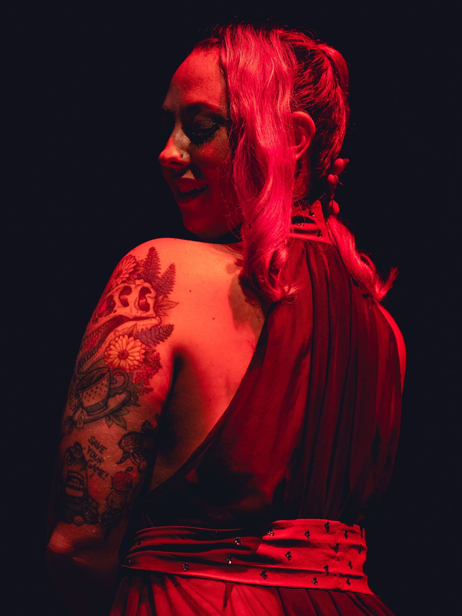 Woman in red backless dress with floral arm tattoo posed under dramatic red lighting.