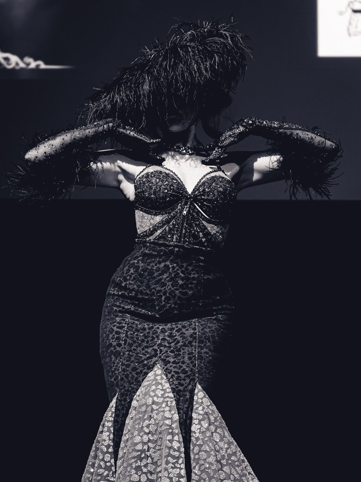Black and white photo of a woman in a sparkly corset gown with feathered headpiece posing dramatically on stage.