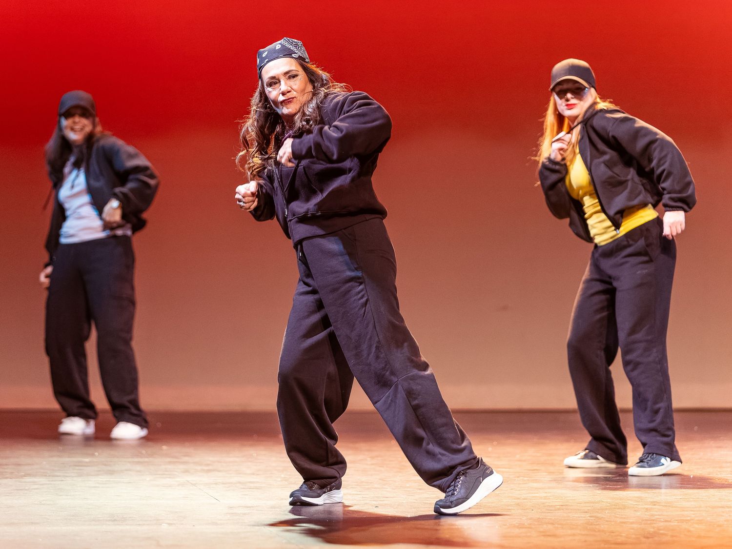 Three female hip-hop dancers perform on stage in matching black outfits with caps under warm red lighting.