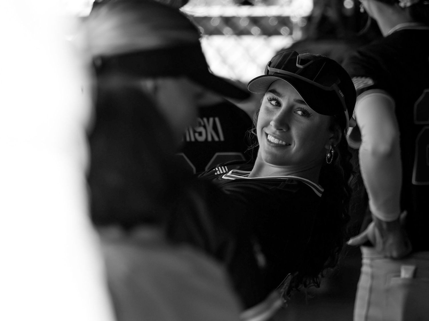 Black and white photo of a smiling woman in a baseball cap enjoying a game in the dugout.